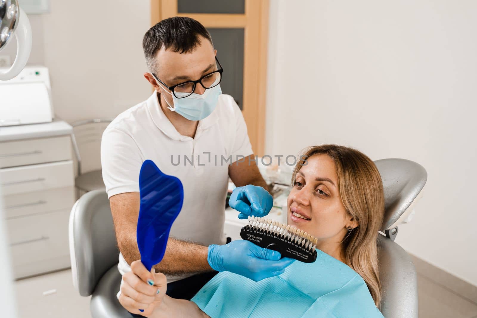 Teeth whitening. Dentist working with teeth color shades guide. Patient looking in mirror. Dentistry. Doctor checking teeth color matching samples in dental clinic. by Rabizo