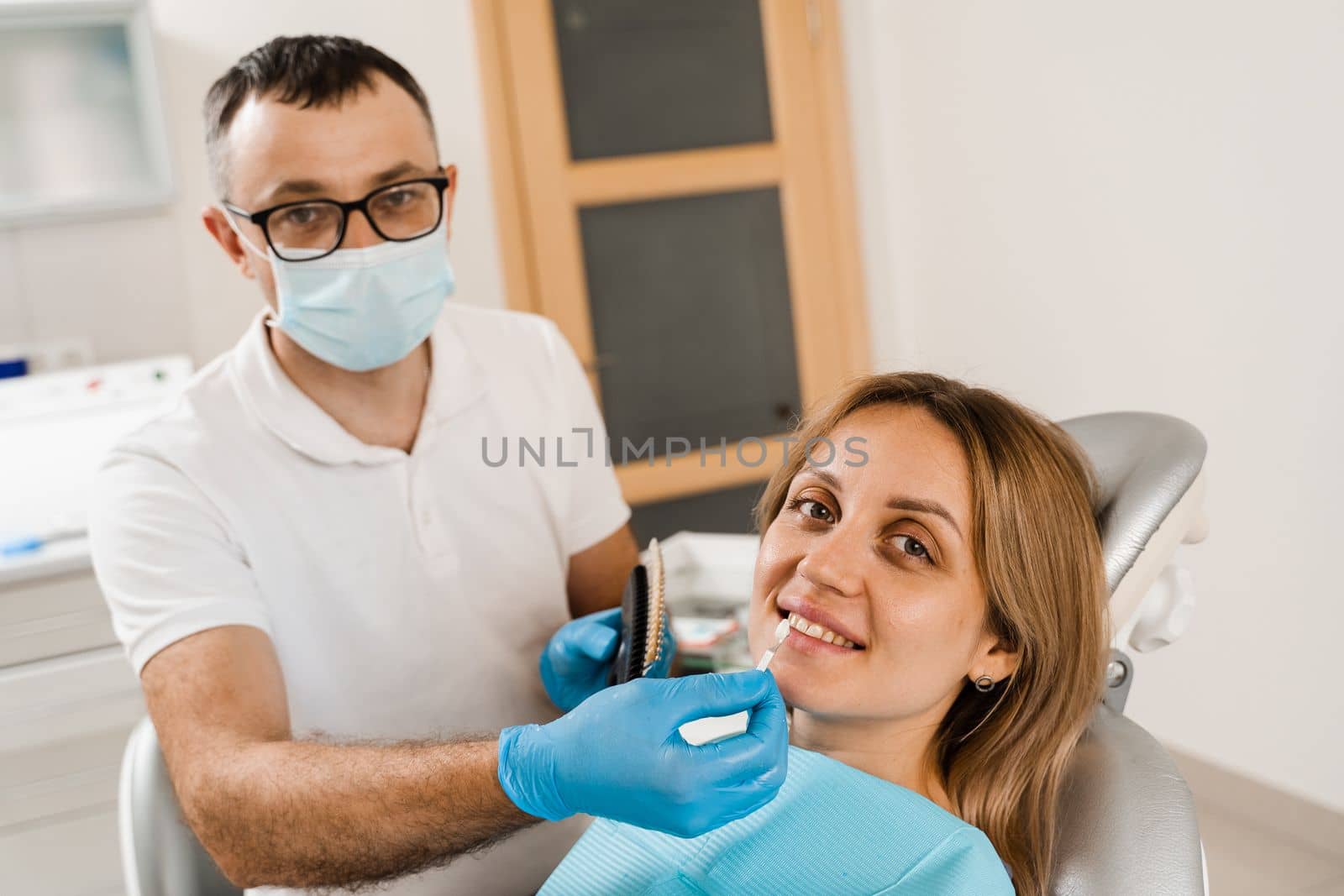 Teeth whitening. Dentist working with teeth color shades guide. Patient looking in mirror. Dentistry. Doctor checking teeth color matching samples in dental clinic. by Rabizo