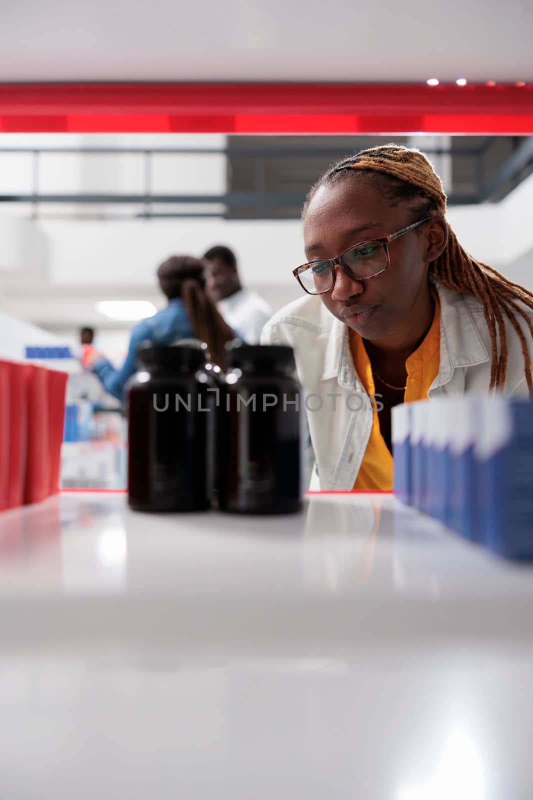 Pharmacy concept, african american pharmacist taking medication bottles from drugstore shelf, selective focus. Customer and seller talking in background, woman putting pills packages close up