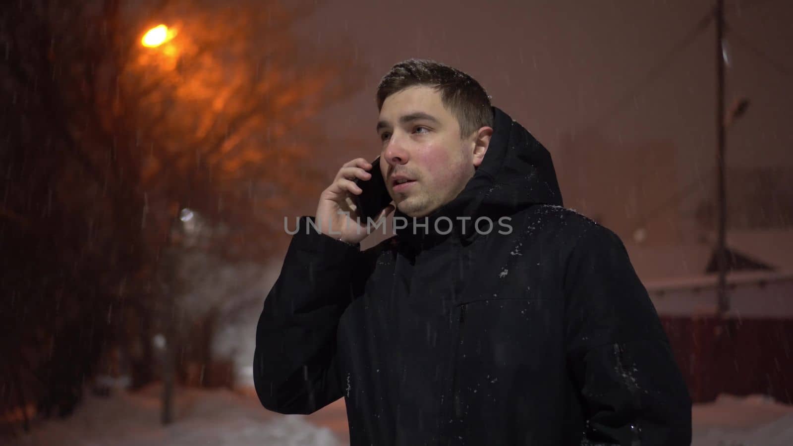 A young man under the snowfall speaks on the phone. Winter warm evening on the street against the background of the light of a lantern. 4k