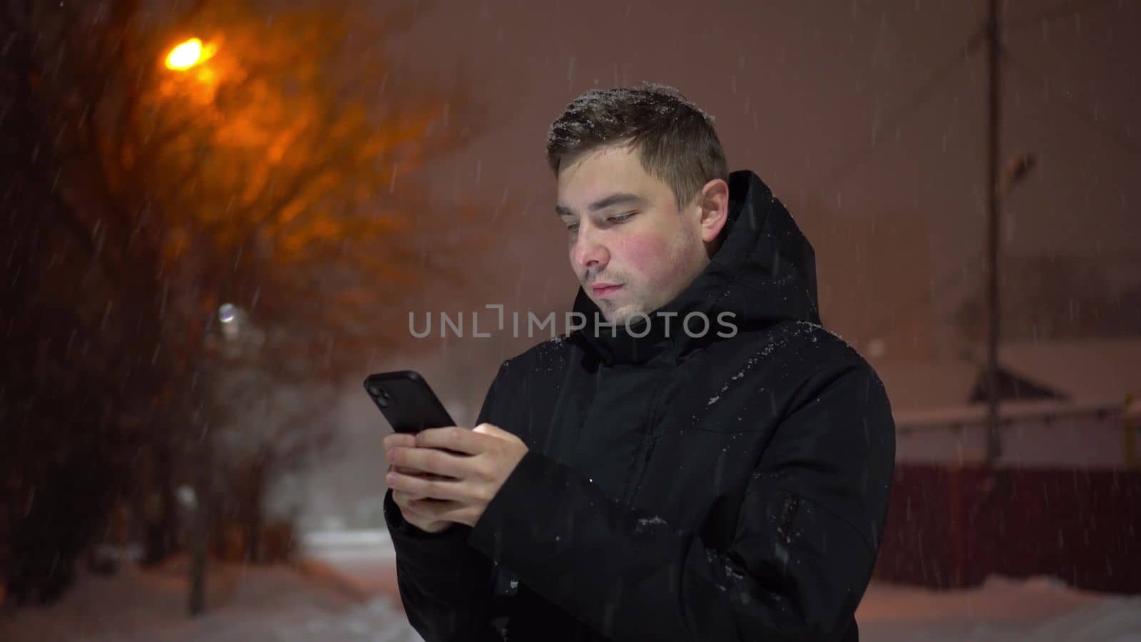 Young man on the street in winter with a phone in his hands. A man under snowfall chatting on a smartphone on a winter evening. 4k