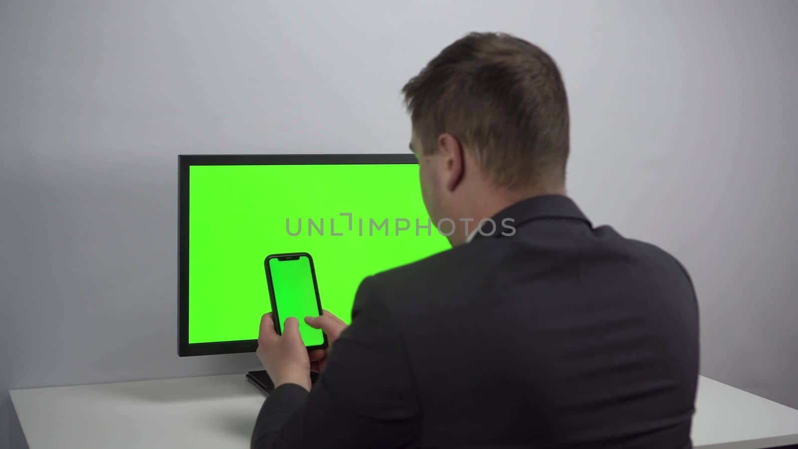 A young businessman sits at a computer and a phone in his hands with green screens. A man works in an office in a jacket. View from the back. 4k