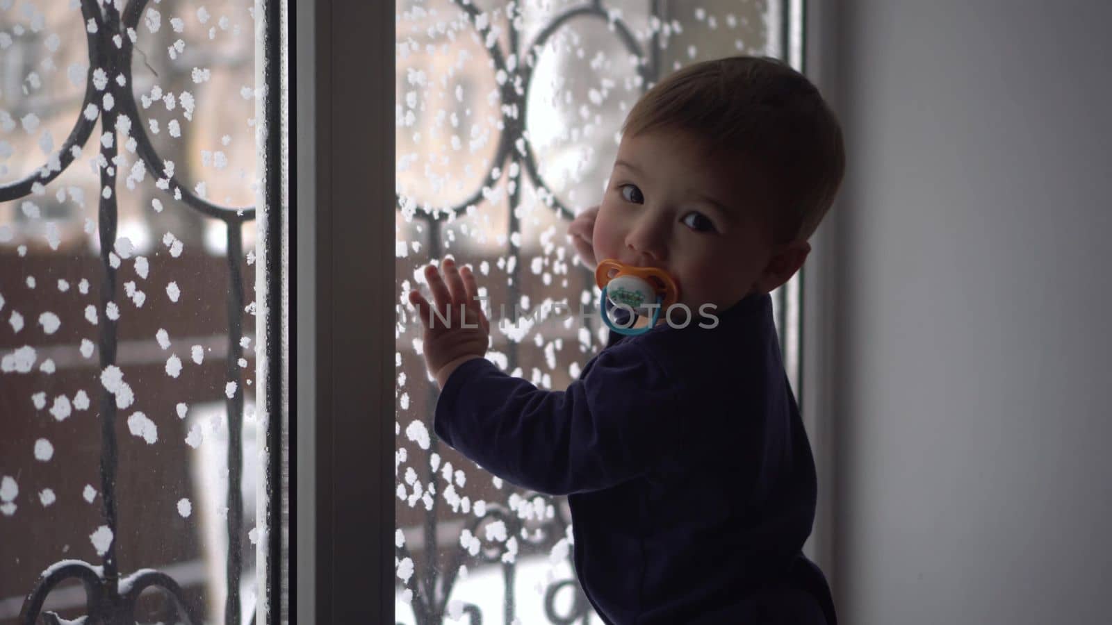 The child looks out the window and knocks on it. A one-year-old son stands at the window in winter. 4k