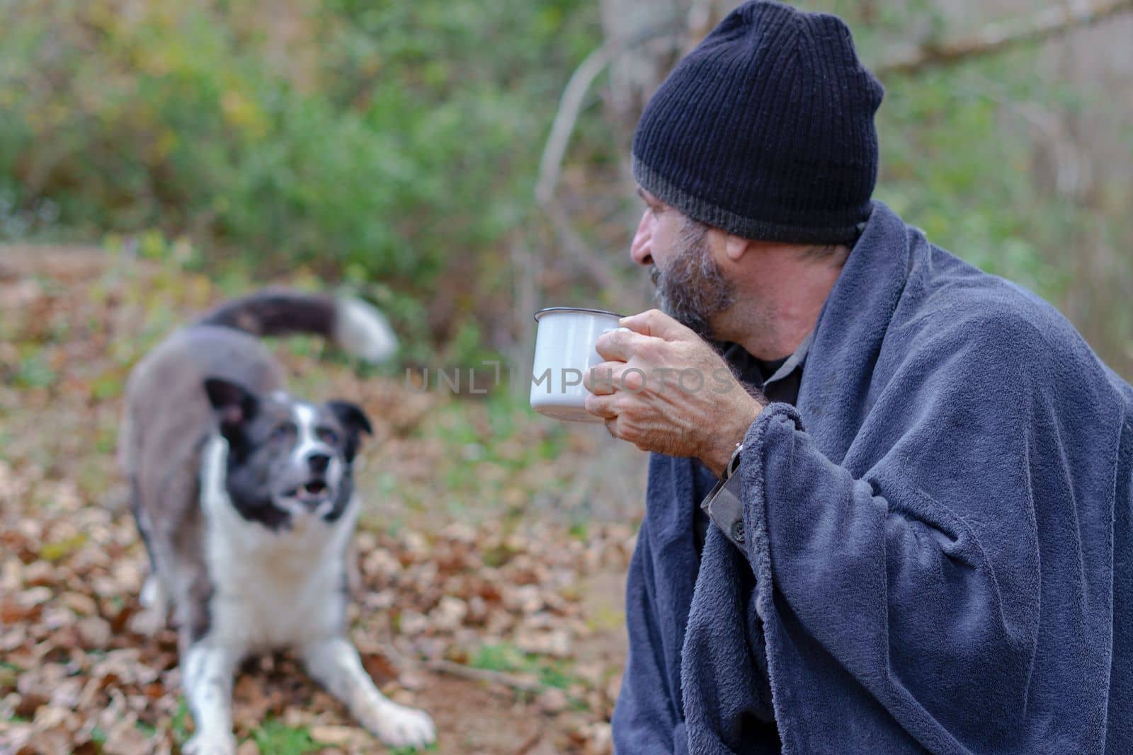 bearded man covered with a blanket drinking a cup of coffee with his dog in the background