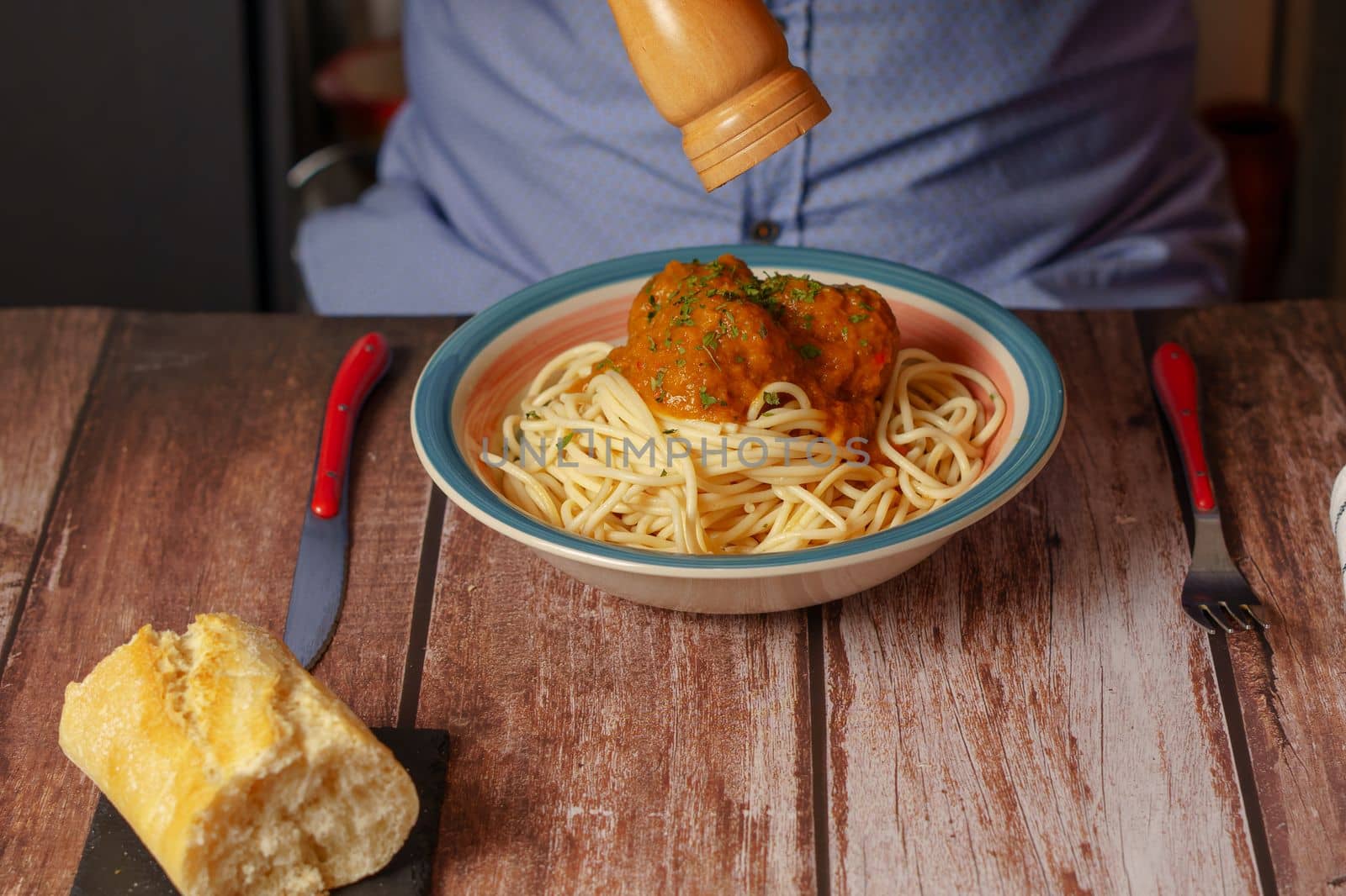 man in blue shirt pouring pepper on a plate of meatballs and pasta with a wooden pepper shaker