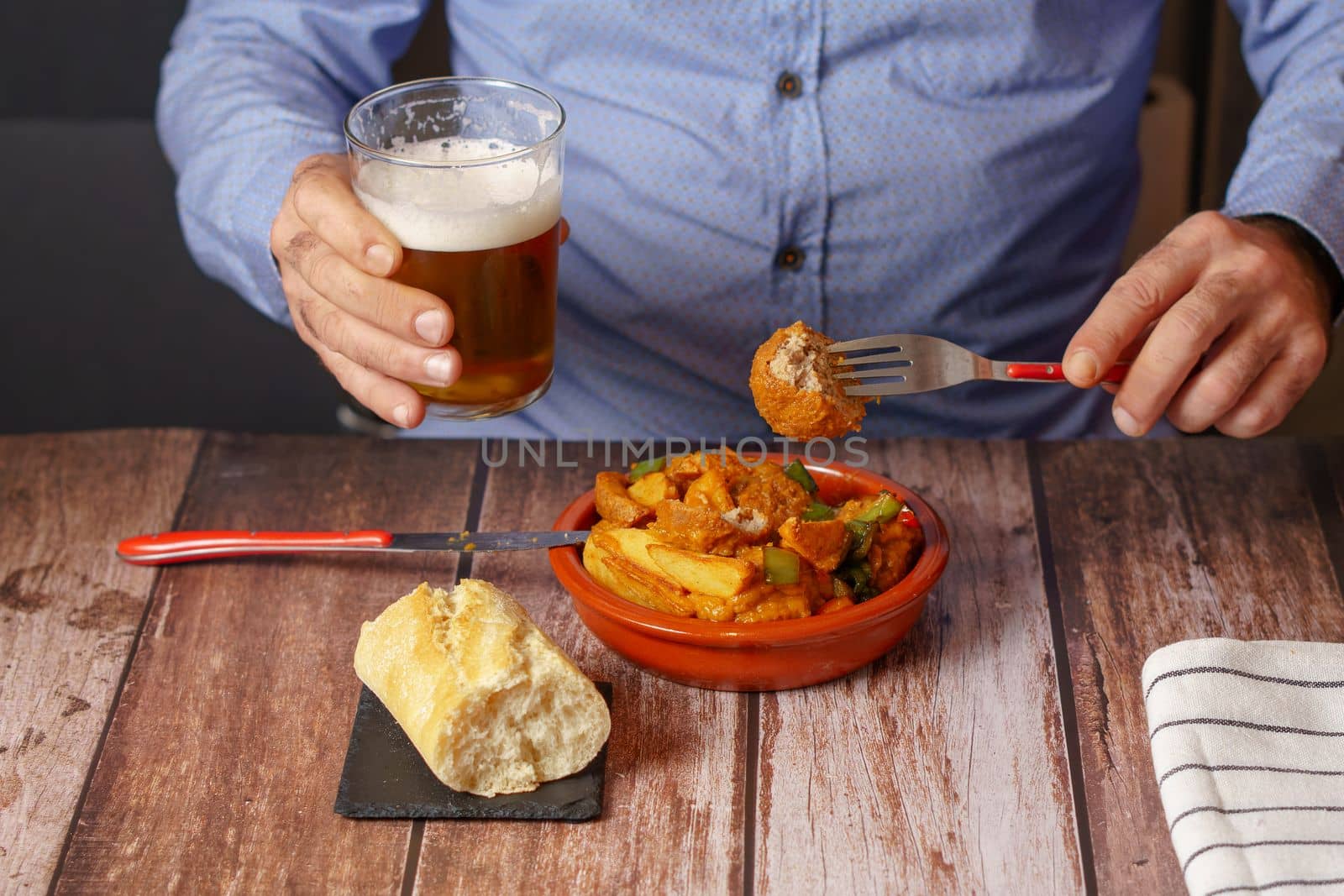man in blue shirt eating a clay pot with meatballs and potatoes and a beer in his hand