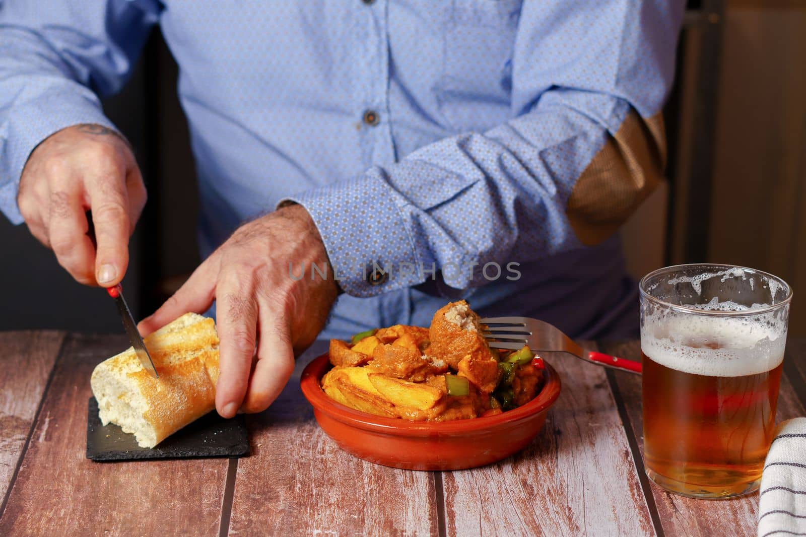 unrecognizable man in blue shirt cutting slice of bread with knife with casserole of meatballs with potatoes and beer