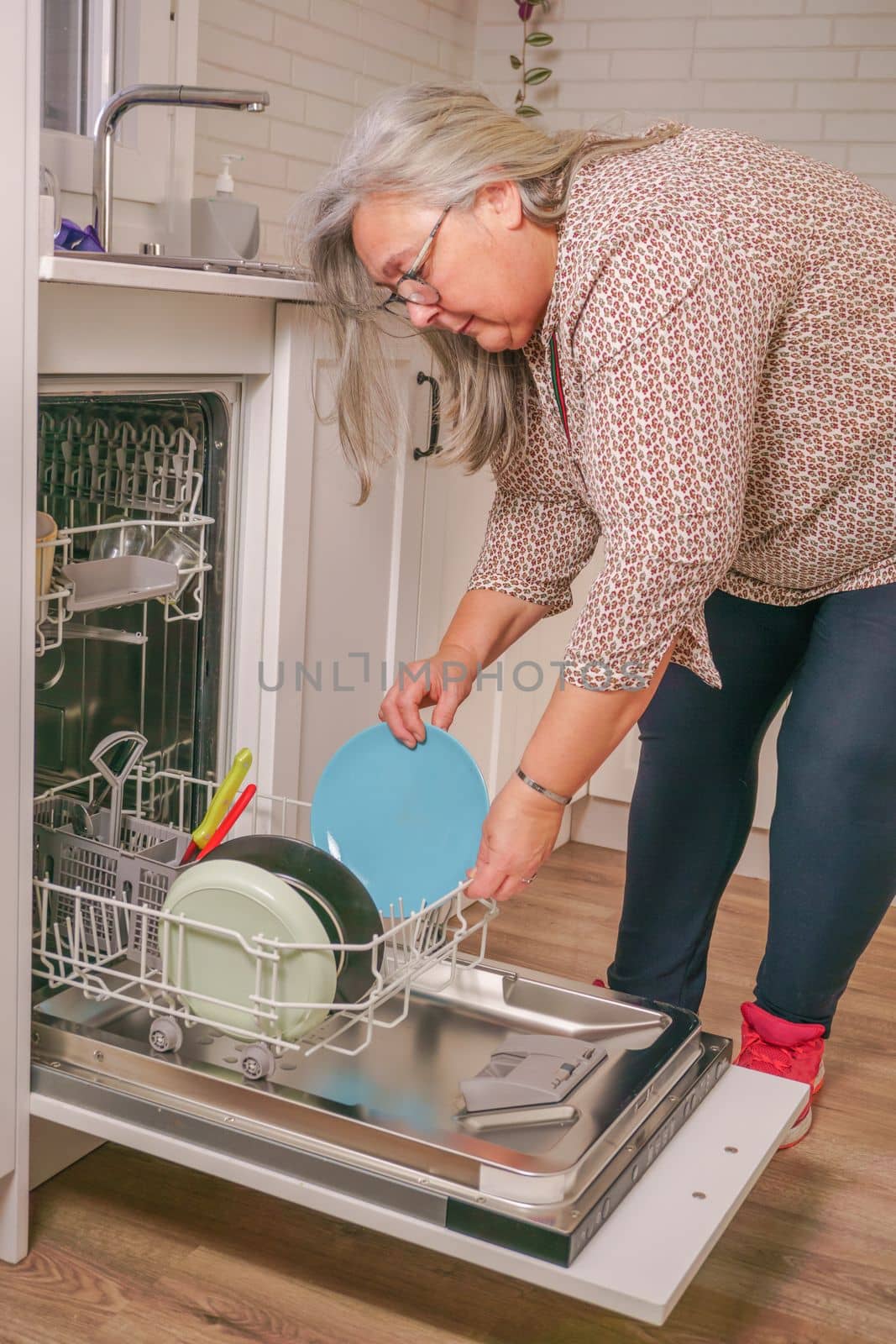 older white-haired woman putting dishes in the dishwasher in her home kitchen