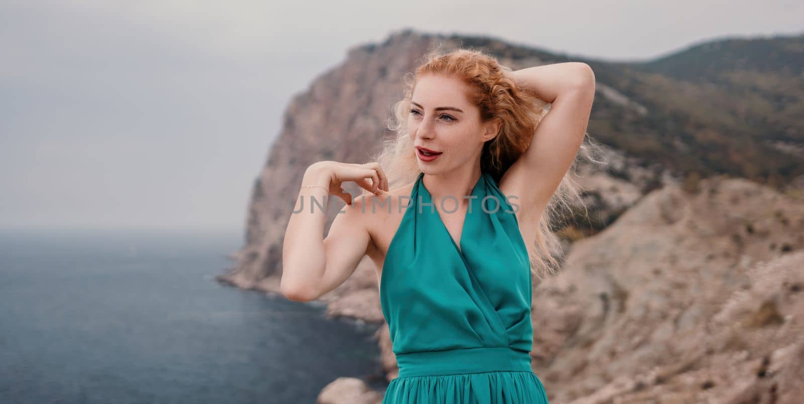 Side view a Young beautiful sensual woman in a mint long dress posing on a volcanic rock high above the sea during sunset. Girl on the nature on overcast sky background. Fashion photo