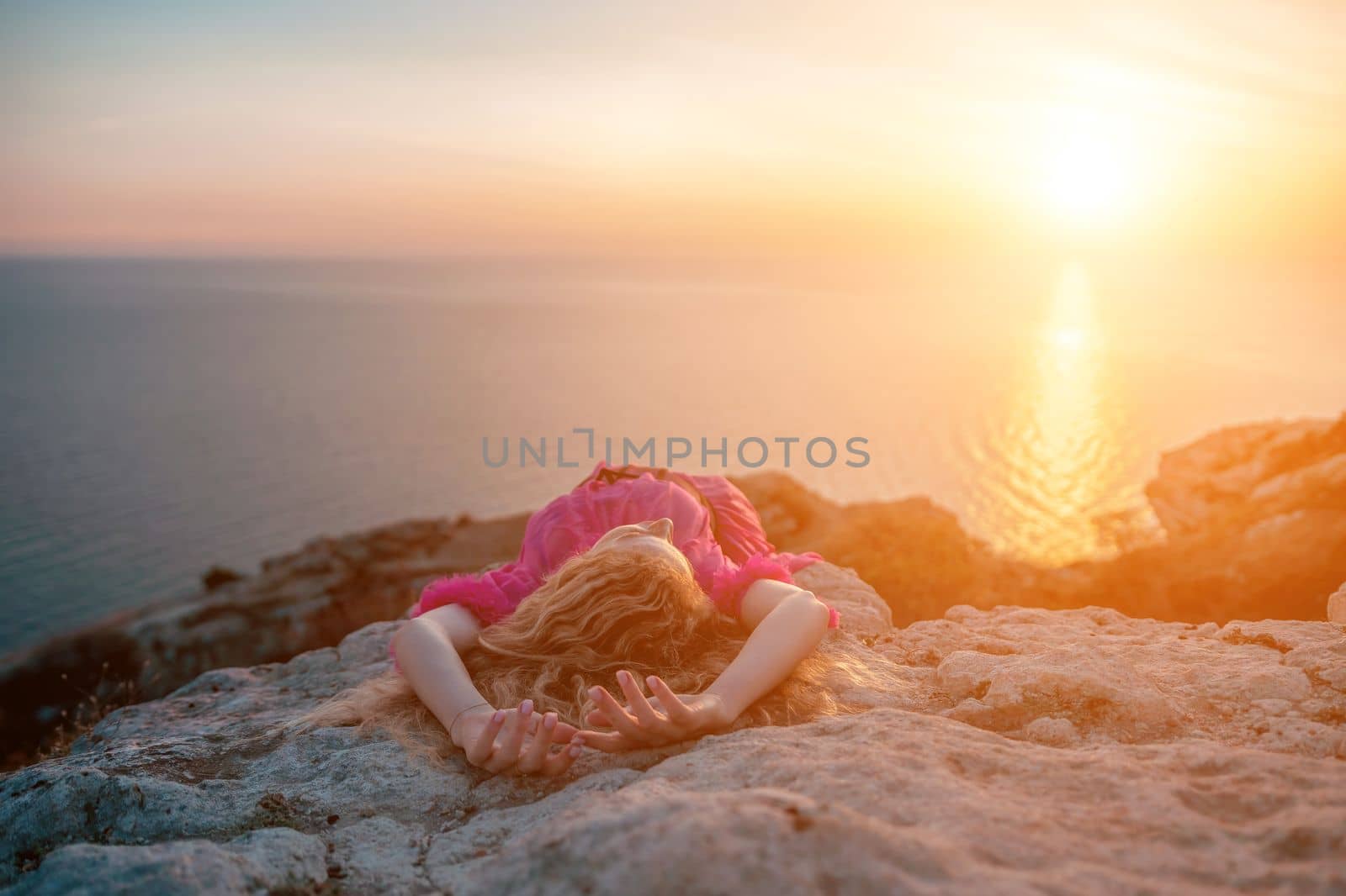 Side view a Young beautiful sensual woman in a red long dress posing on a volcanic rock high above the sea during sunset. Girl on the nature on blue sky background. Fashion photo