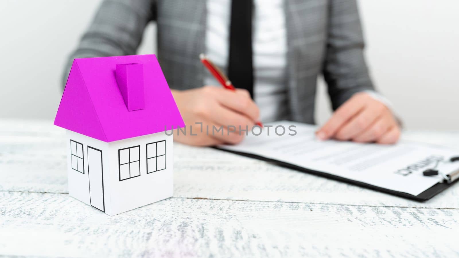 Businesswoman sitting and writing in notebook. Paper house on desk.
