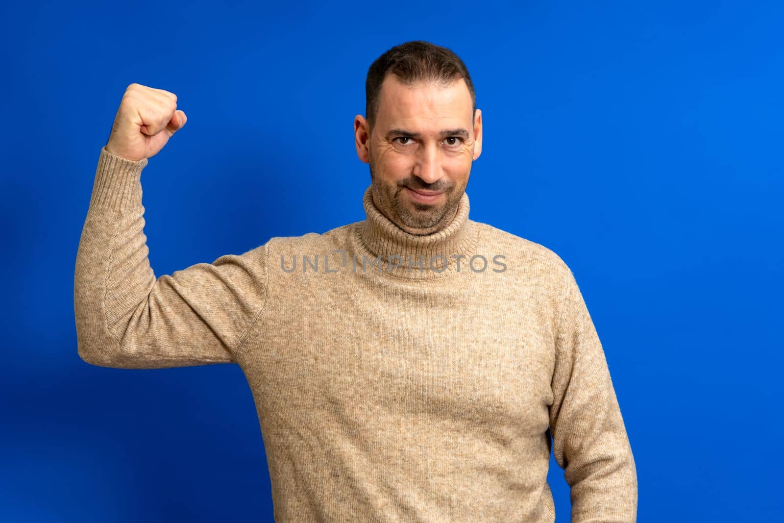 Latino male happy unshaven strong 40s man in beige turtleneck showing biceps muscles on hand isolated on blue color background studio portrait