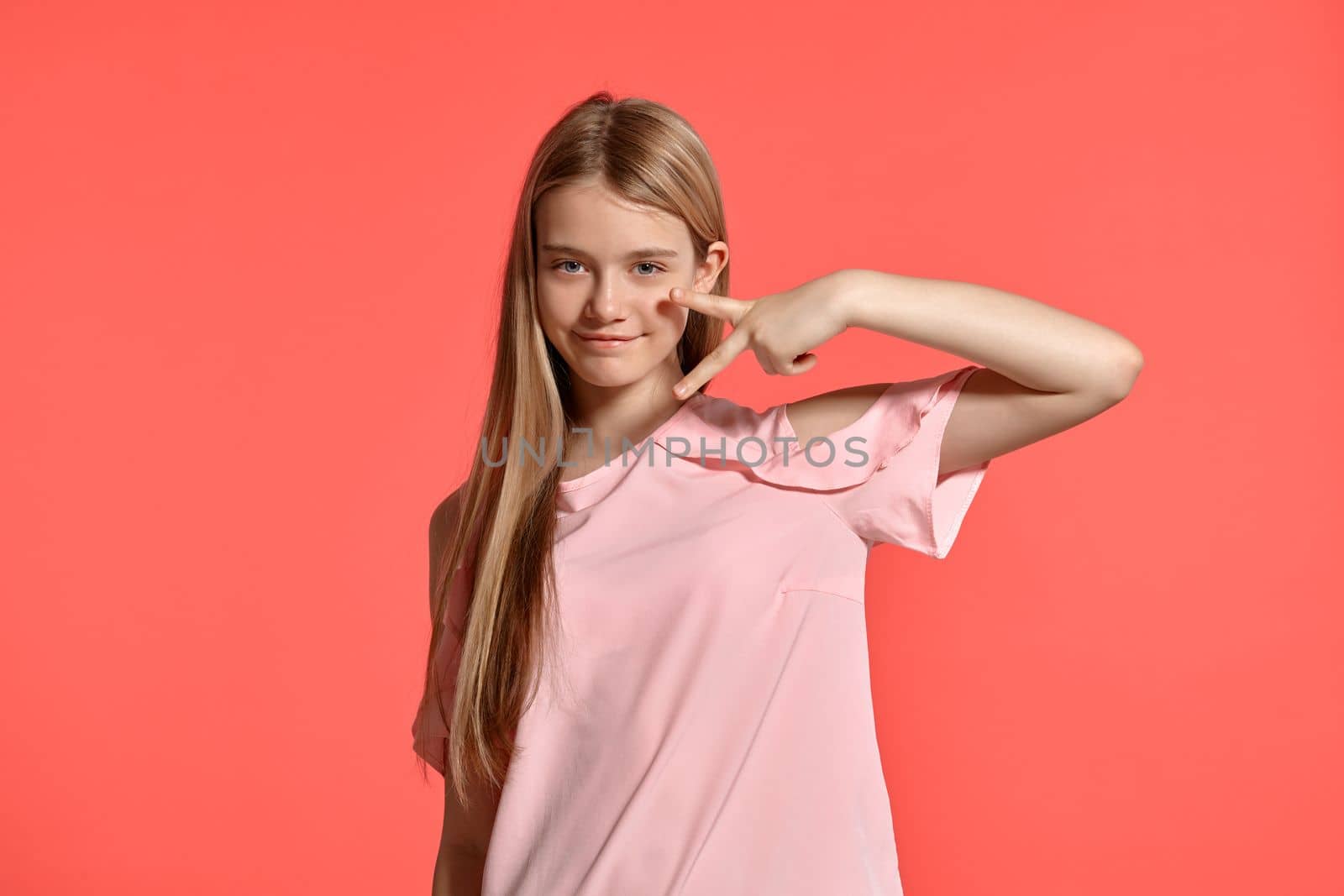 Studio portrait of a nice blonde teenage girl with a long hair, in a rosy t-shirt, standing against a pink background in various poses. She expresses different emotions posing sideways right in front of the camera, smiling and showing a peace sign.