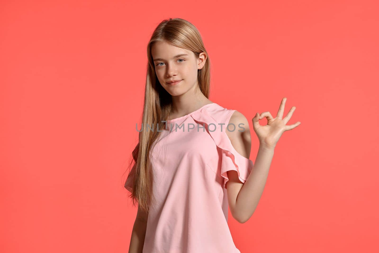 Studio portrait of a beautiful blonde teenager with a long hair, in a rosy t-shirt, standing against a pink background in various poses. She expresses different emotions posing right in front of the camera, smiling and showing ok sign.