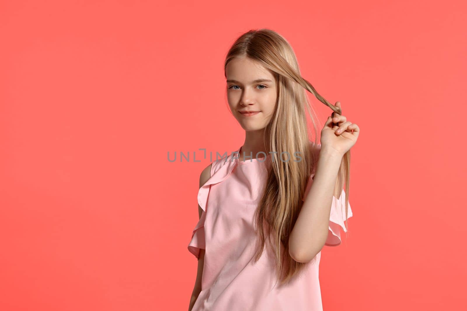 Studio portrait of a beautiful blonde teenage girl with a long hair, in a rosy t-shirt, standing against a pink background in various poses. She expresses different emotions posing right in front of the camera, smiling and winding hair on a finger.