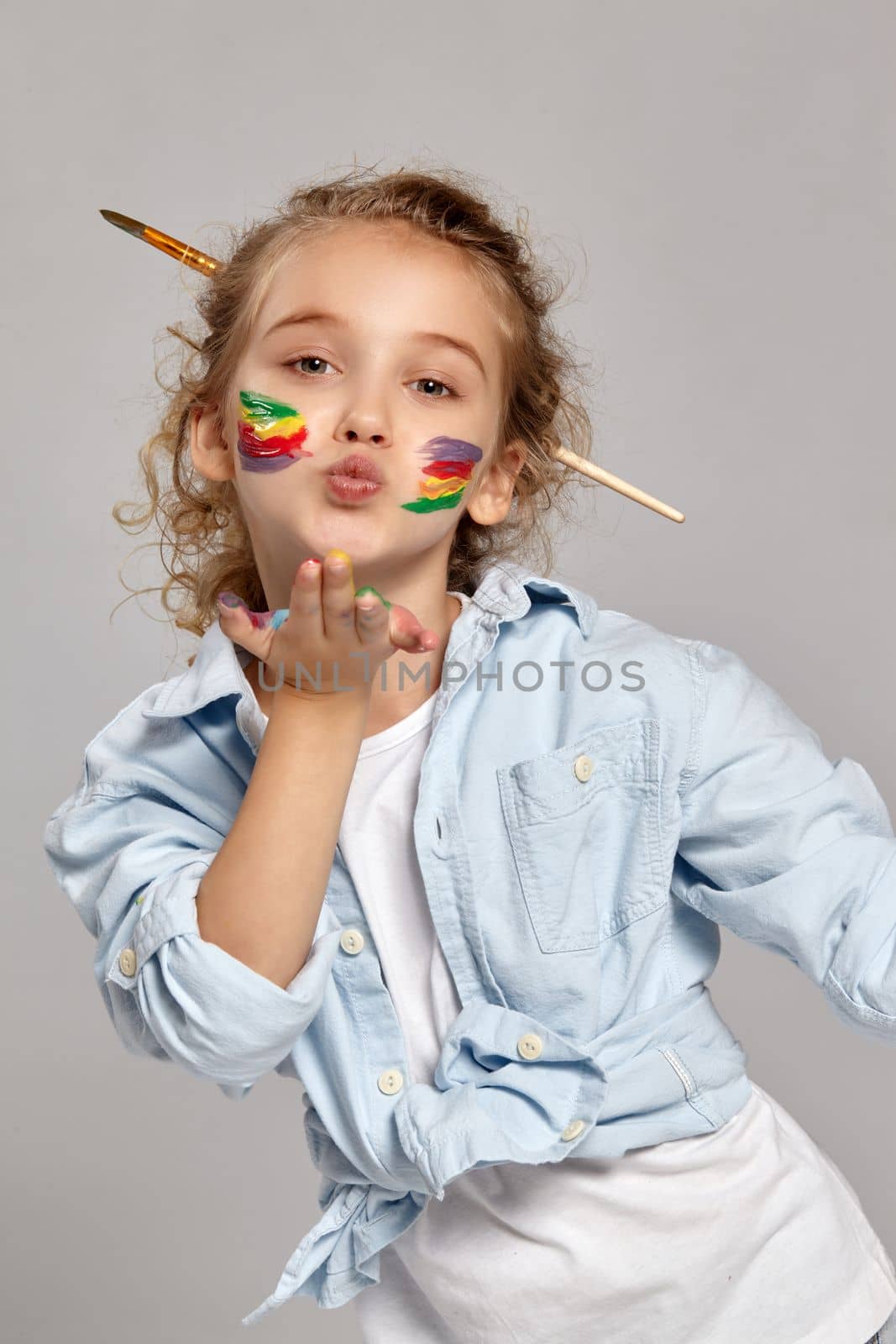 Charming girl with a painted cheeks having a brush in her chic curly blond hair, wearing in a blue shirt and white t-shirt. She is giving a kiss at the camera, her arms are painted too, on a gray background.