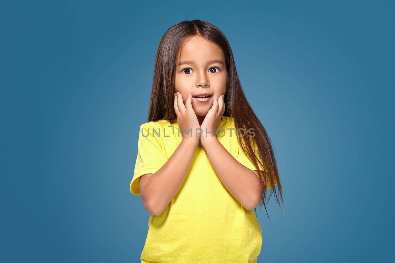 Close up portrait of cute joyful pretty littlegirl with excellent skin and beaming smile, she is admiring her beauty in a mirror, on blue background