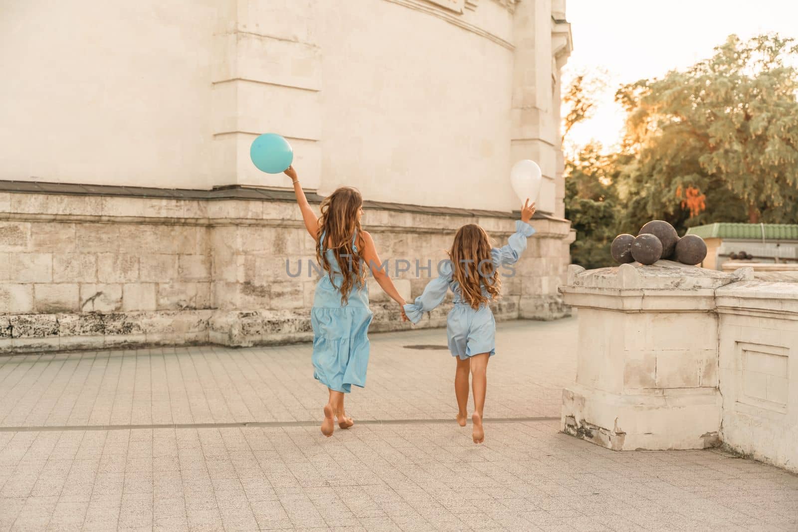 Daughter mother run holding hands. In blue dresses with flowing long hair, they hold balloons in their hands against the backdrop of a sunset and a white building