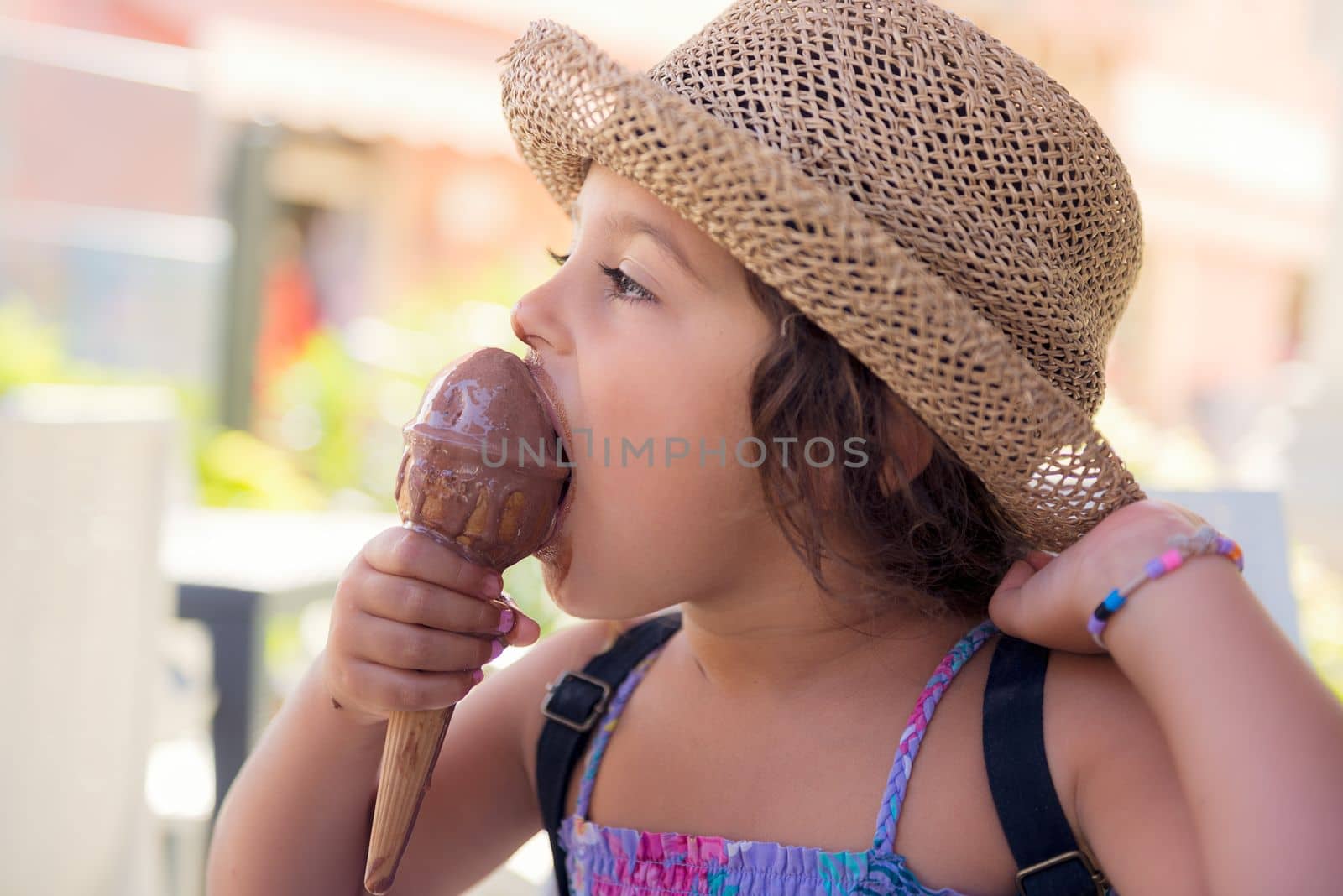 Little girl with a straw hat and a summer dress enjoys the summer heat eating a refreshing ice cream cone, it melts in her hand while she smiles funny with her mouth and nose stained with chocolate