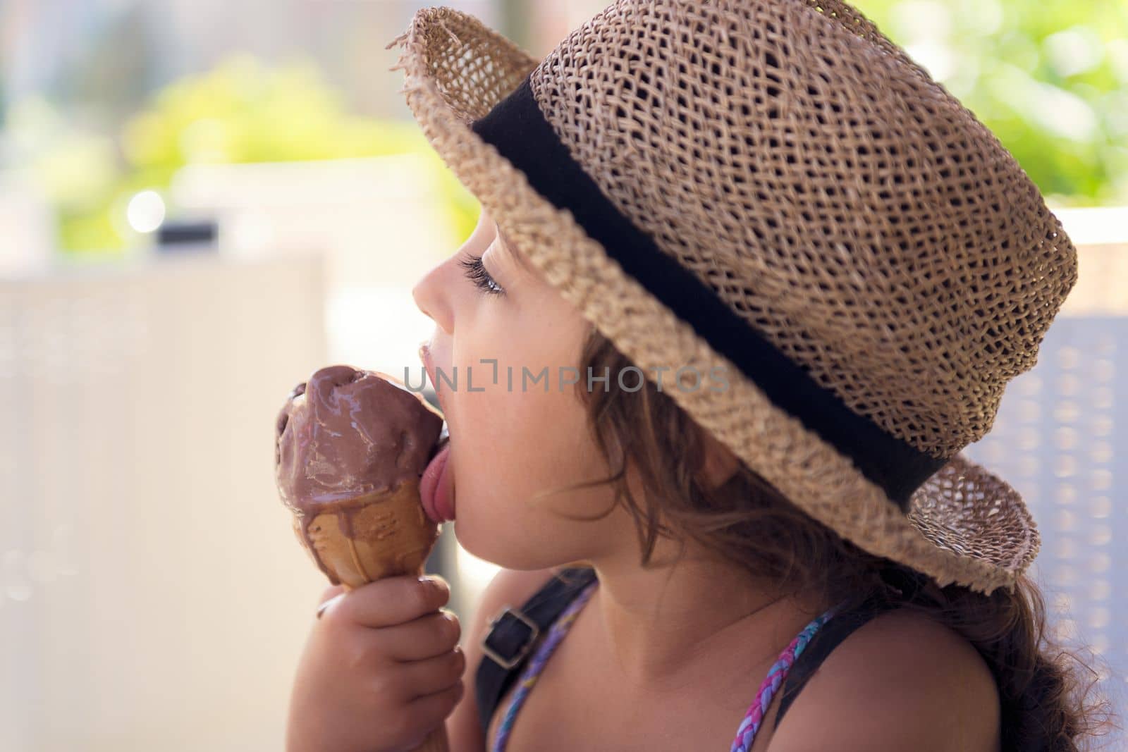 Little girl with a straw hat and a summer dress enjoys the summer heat eating a refreshing cone of chocolate ice cream, it melts in her hand while she licks it with her tongue