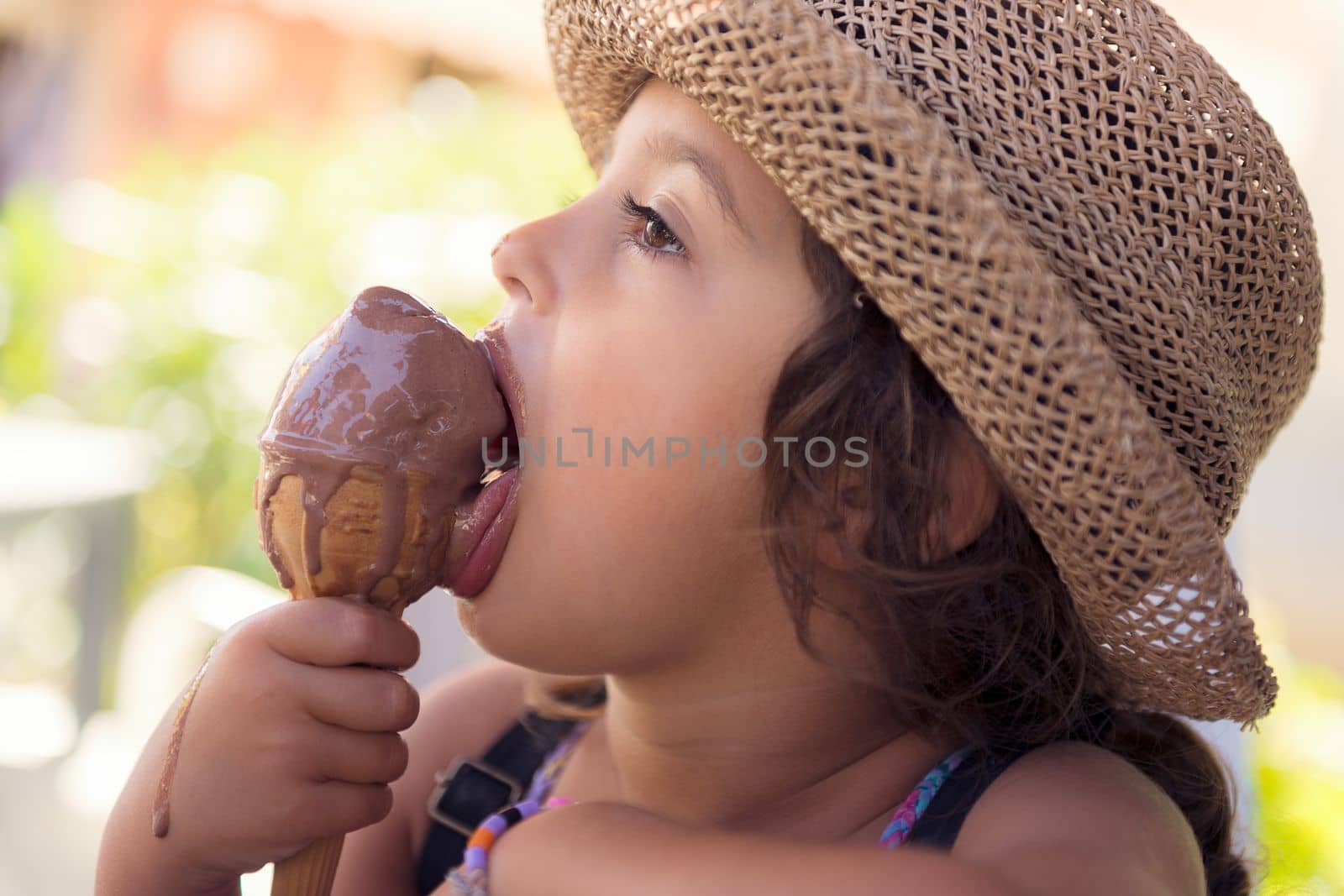 Little girl with a straw hat and a summer dress enjoys the summer heat eating a refreshing cone of chocolate ice cream, it creamy melts in her hand while she licks it with her tongue