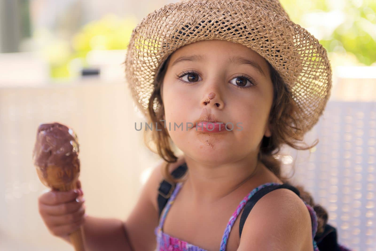 Little girl with straw hat and a sundress enjoys the summer heat eating a refreshing cone of chocolate ice cream, it melts on her hand and she has the nose stained with chocolate
