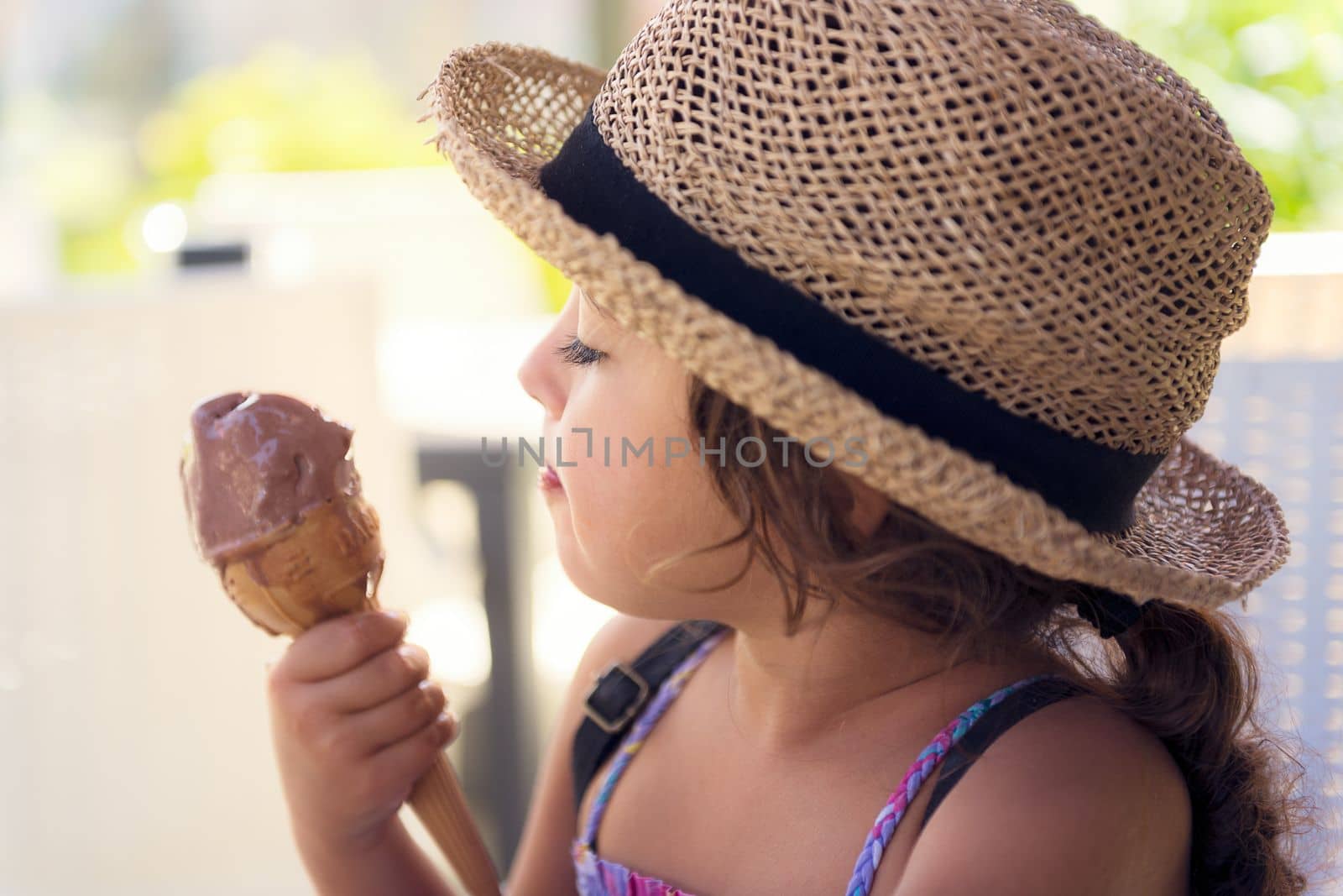 Little girl with a straw hat and a summer dress enjoys the summer heat eating a refreshing cone of chocolate ice cream, it melts in her hand while she looks at it with desire
