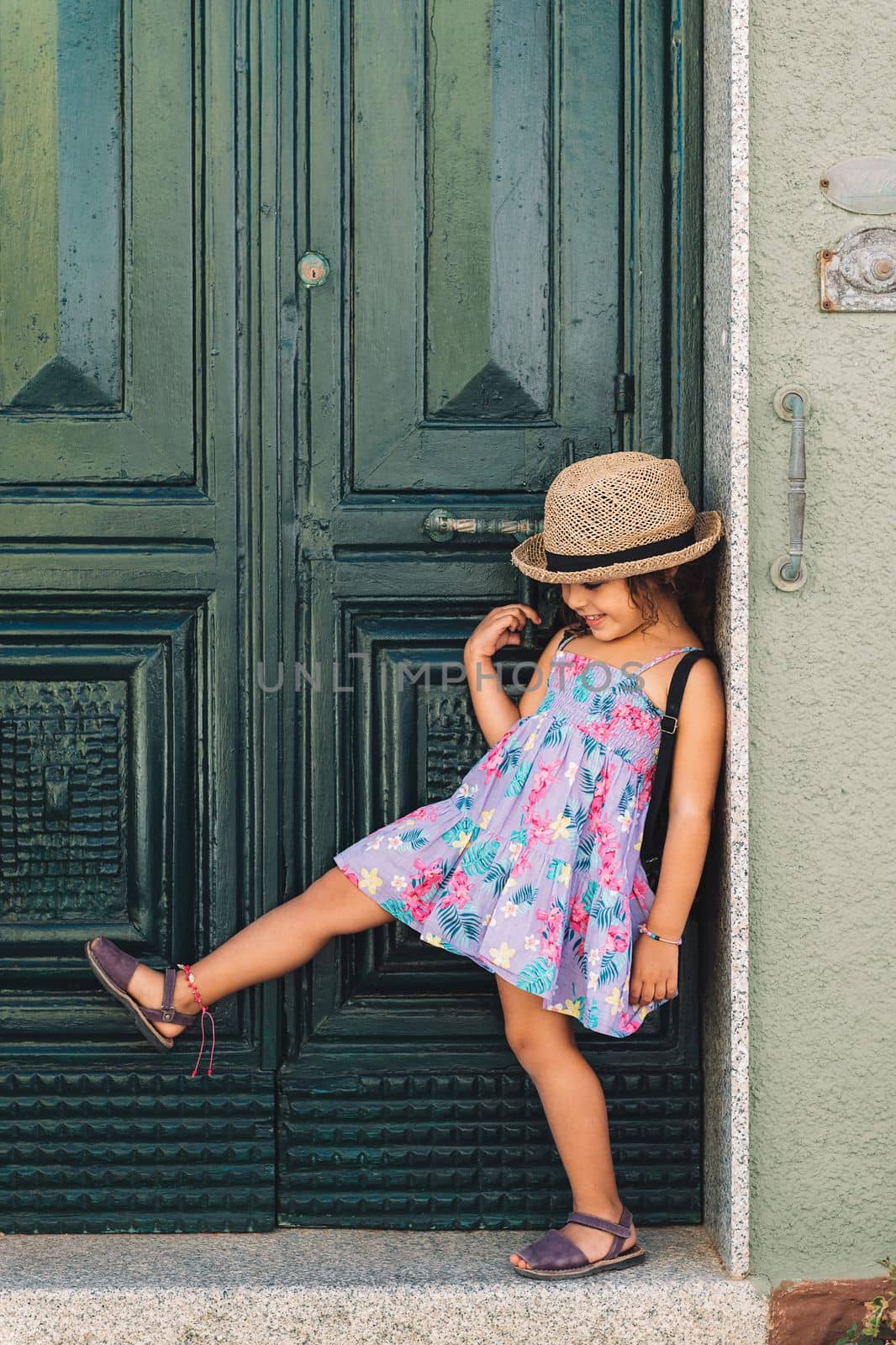 Smiling little girl lifts a leg next to the green door of her house, she wears a lilac summer dress of flowers and a, vertical image