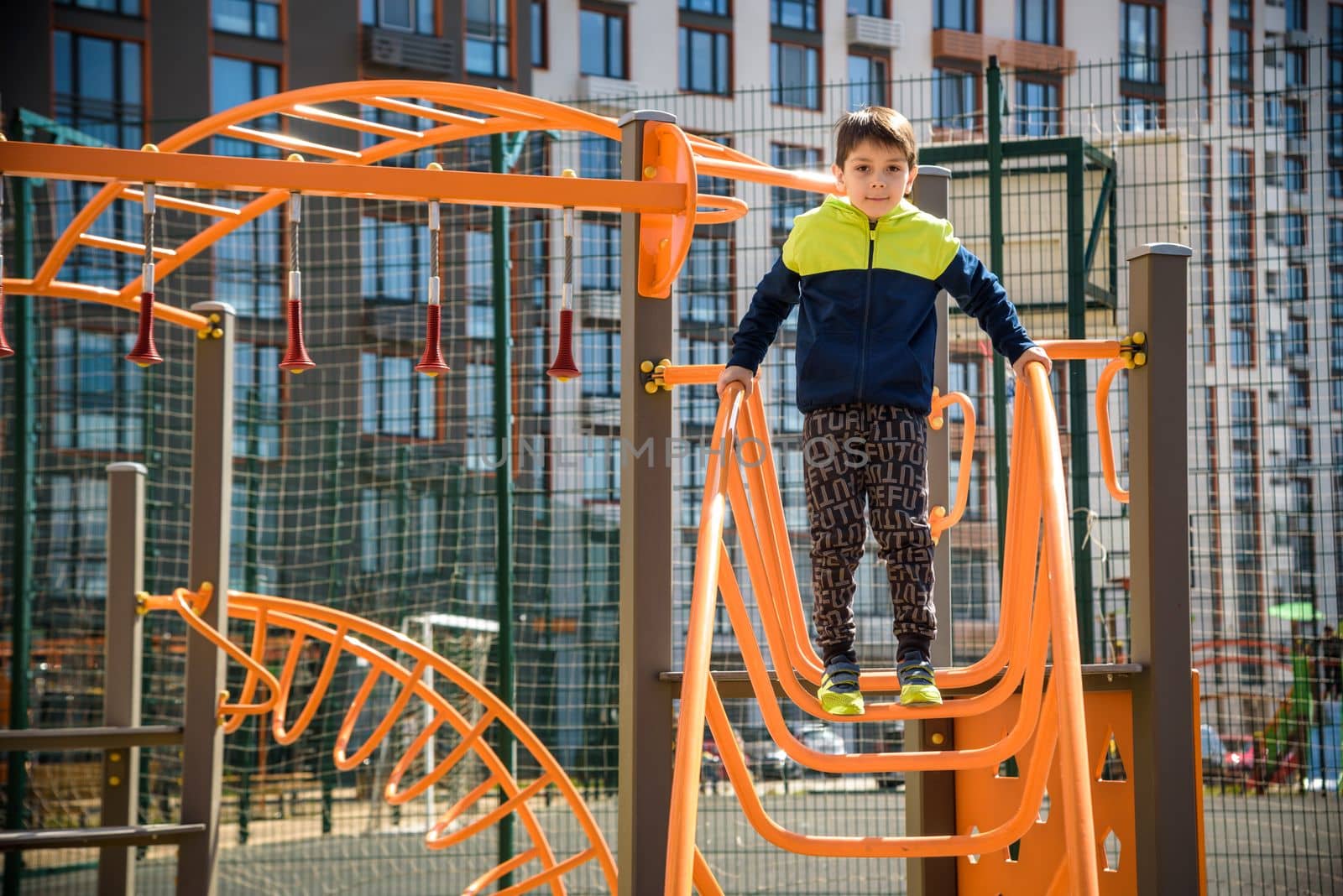 Cute boy is climbing on the playground in the schoolyard. He has a very happy face and enjoy this adventure sports alone outdoor. Warm sunny day by Kobysh