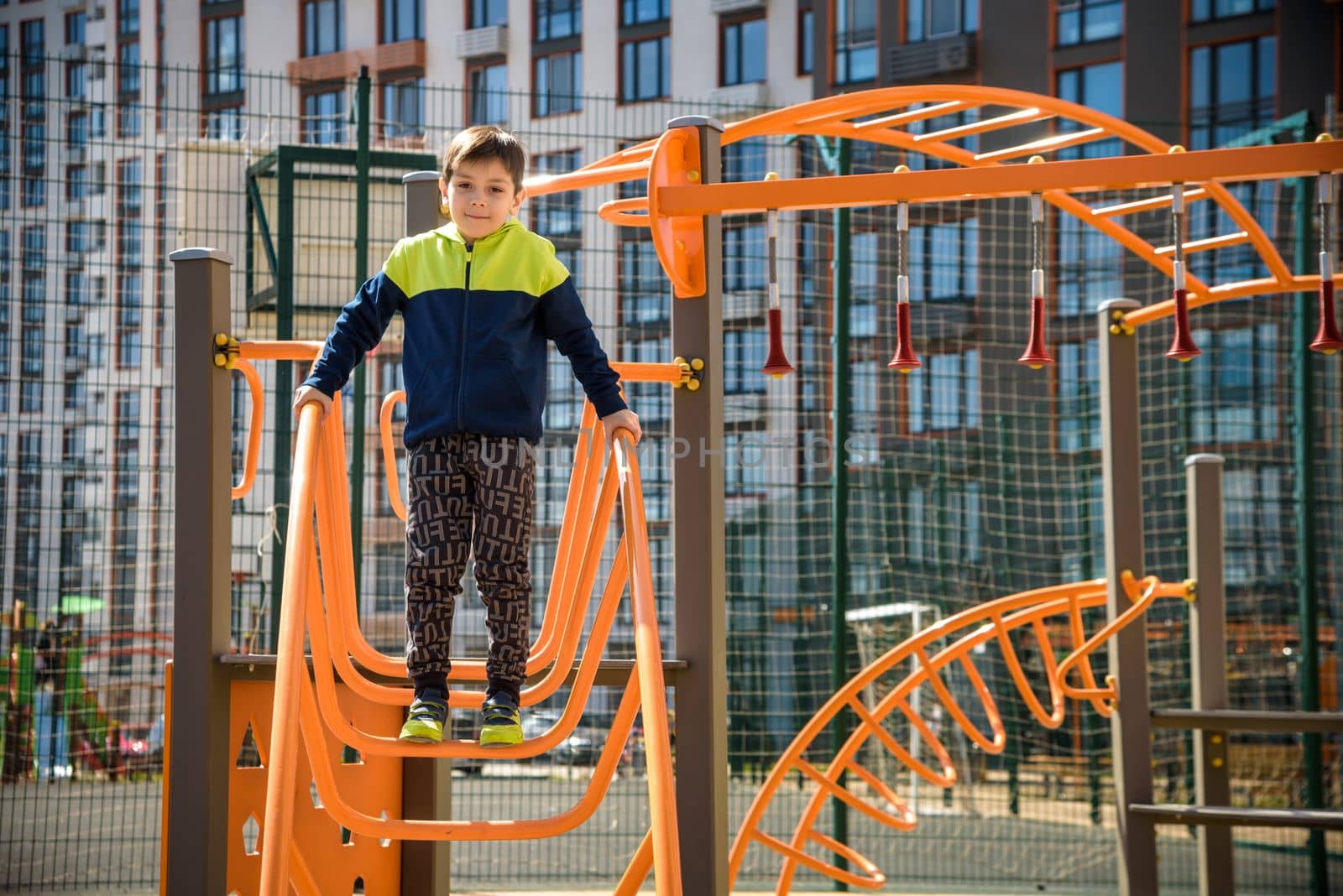 Cute boy is climbing on the playground in the schoolyard. He has a very happy face and enjoy this adventure sports alone outdoor. Warm sunny day by Kobysh