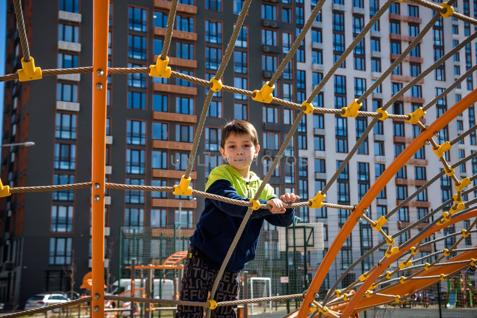 Cute boy is climbing on the playground in the schoolyard. He has a very happy face and enjoy this adventure sports alone outdoor. Warm sunny day by Kobysh