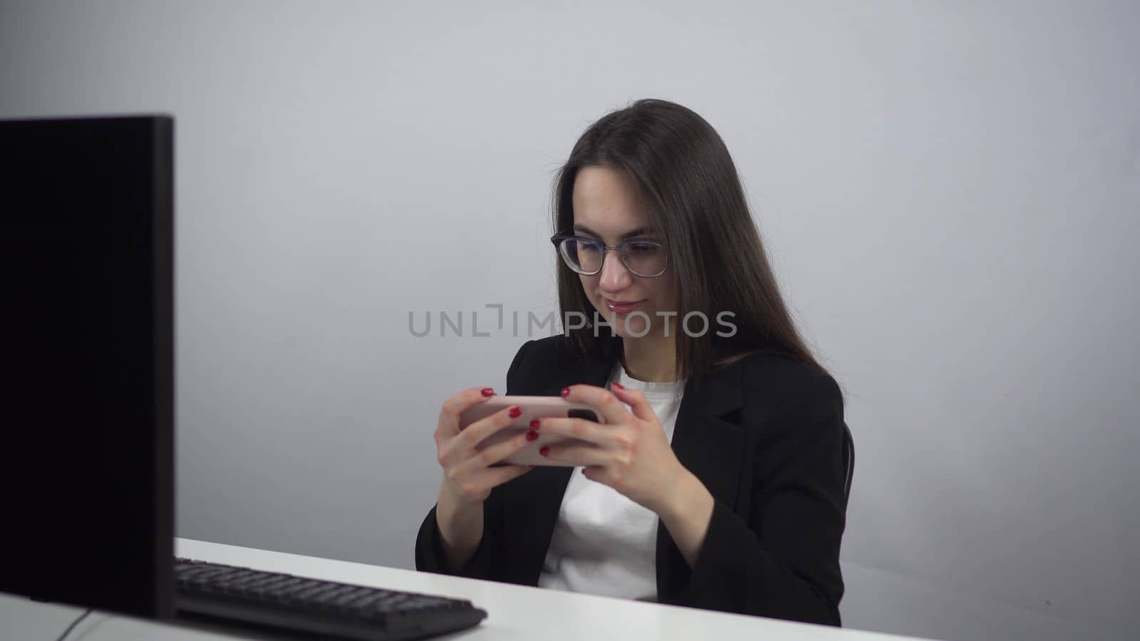 Businesswoman plays a mobile game in the office. A young woman in a jacket and glasses plays on a smartphone while working. 4k