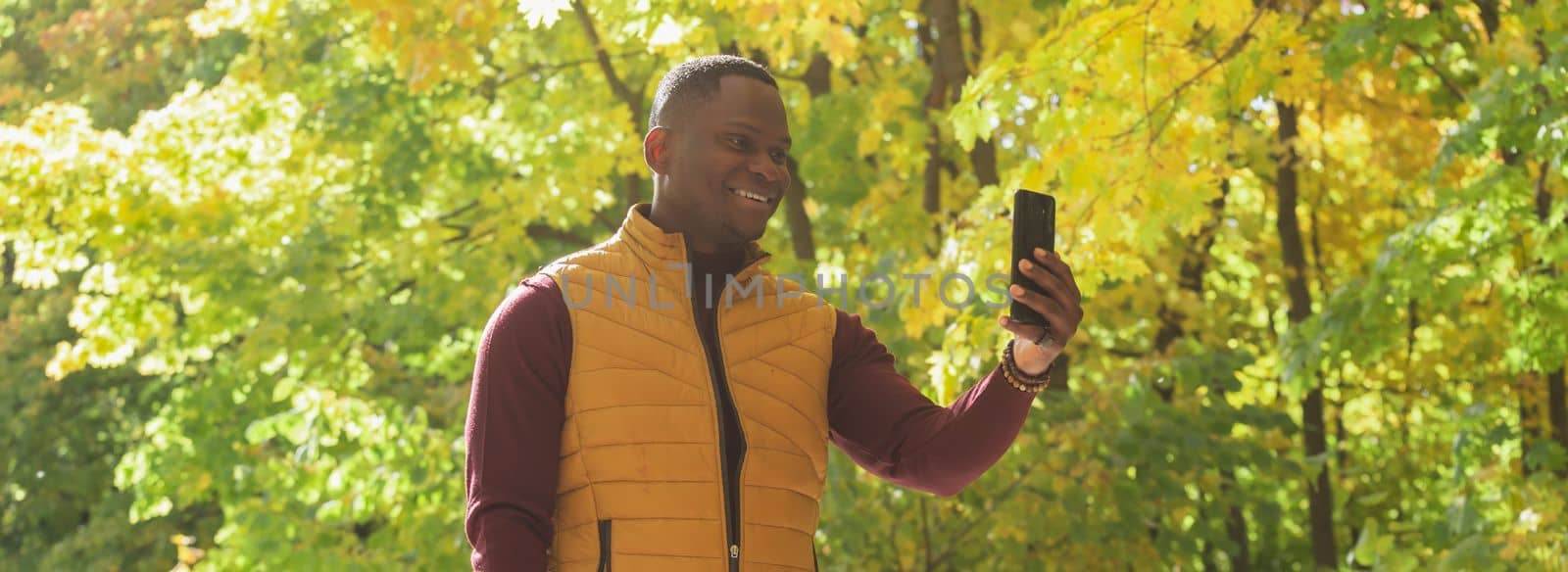 Banner african American man taking a self portrait with a smartphone in autumn fall park copy space and place for text. Happy people, season and new normality concept by Satura86
