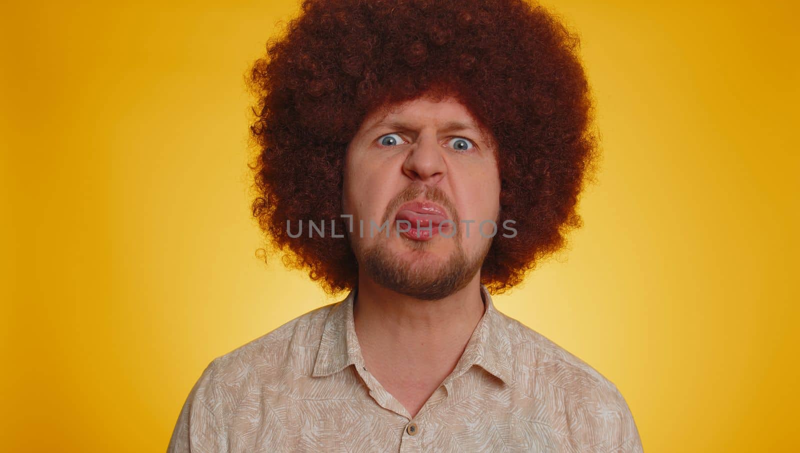 Funny joyful sincere man with lush Afro hairstyle coiffure making playful silly facial expressions and grimacing fooling around showing tongue. Young handsome hipster guy isolated on yellow background