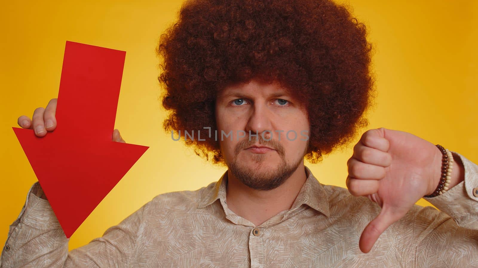 Upset man with afro hairstyle showing red arrow pointing down, concept of downgrade, unsuccessful business, fall of stock market money exchange rate, bankruptcy fail. Hipster guy on yellow background
