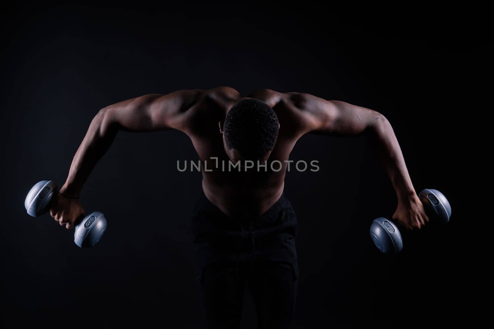 Confident young man shirtless portrait training with dumb-bell against black background.