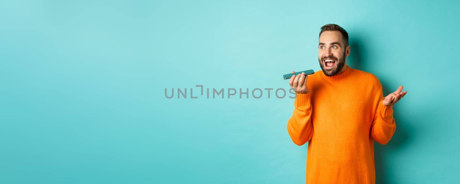 Happy man talking on speakerphone, gesturing and recording voice message on mobile phone, standing in orange sweater over light blue background.