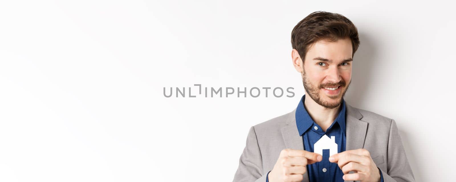 Real estate and insurance concept. Smiling male realtor showing small paper house cutout and looking at camera, selling property, white background.
