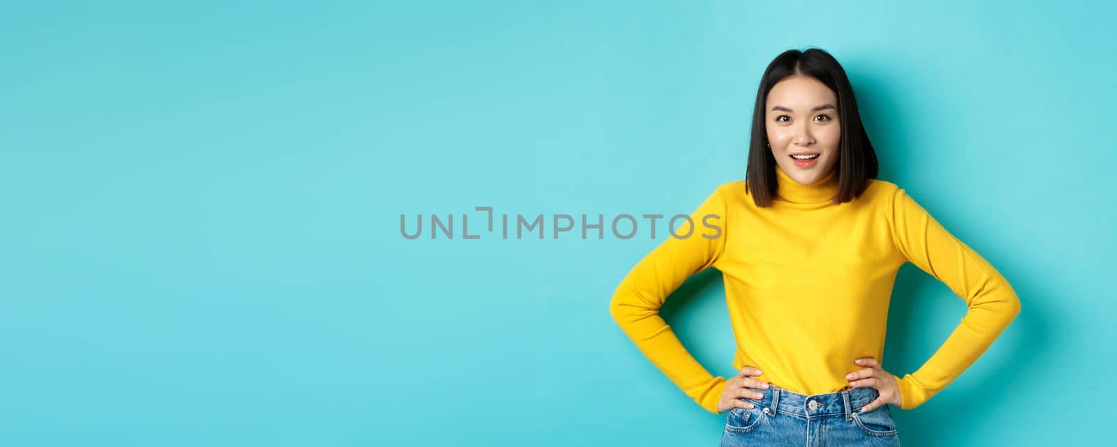 Image of impressed korean girl listening interesting story, looking at camera intrigued, standing over blue background.