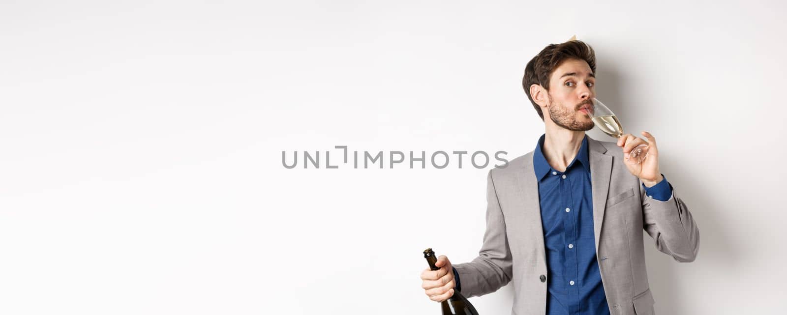 Celebration and holidays concept. Handsome bearded man in suit and birthday hat holding bottle, drinking glass of champagne, standing on white background by Benzoix