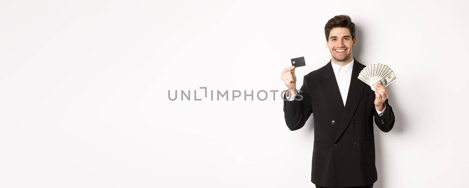 Portrait of attractive businessman in black suit, showing money and credit card, smiling pleased, standing against white background.