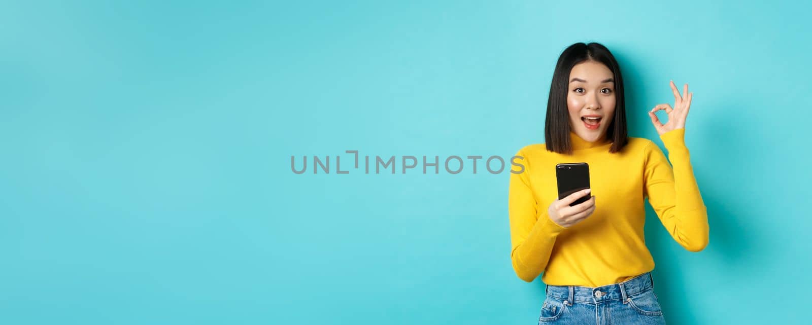 E-commerce and online shopping concept. Portrait of asian woman showing OK sign and using mobile phone, recommend application, standing over blue background.