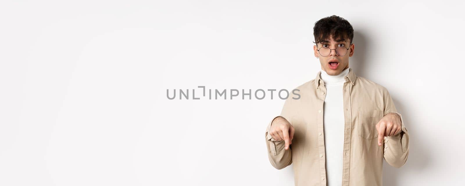 Shocked attractive young man showing something awesome, drop jaw and gasping in awe, pointing fingers down, standing on white background.