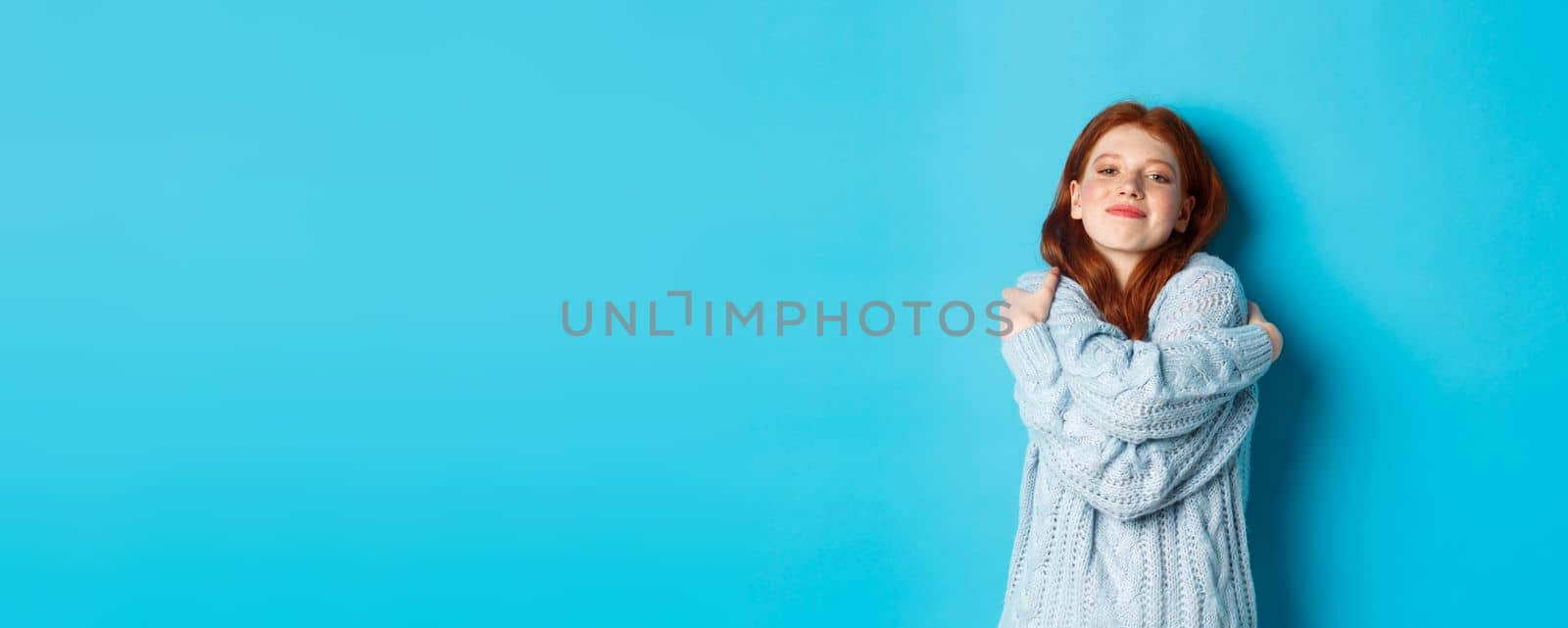Happy cute redhead girl hugging herself, wearing comfortable and warm sweater, smiling at camera, standing over blue background.