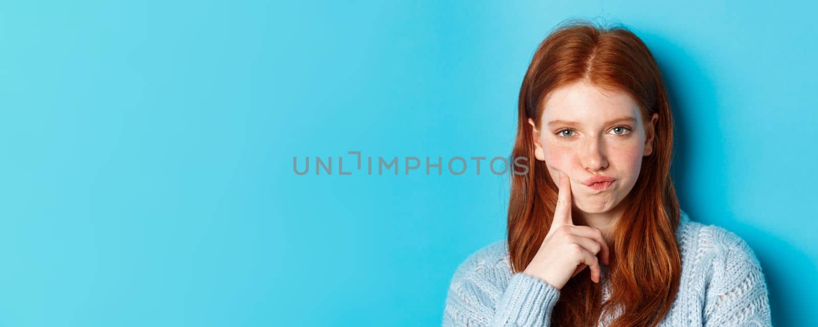 Puzzled redhead girl looking suspicious at camera, thinking or solving problem, standing against blue background.