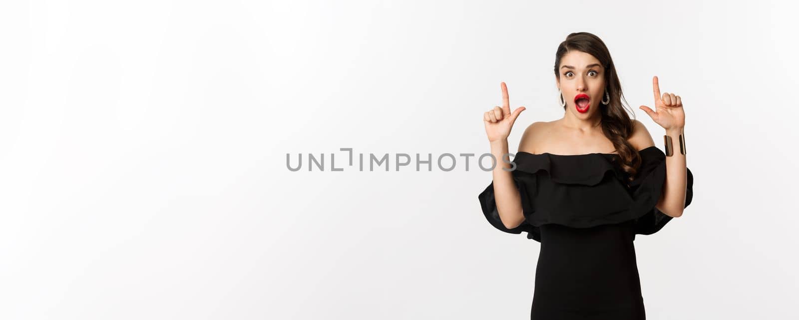 Fashion and beauty. Surprised woman in black dress pointing fingers up, showing banner, standing over white background by Benzoix