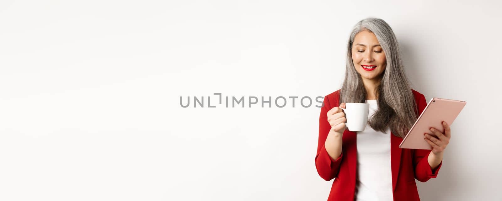 Business people concept. Asian mature businesswoman holding digital tablet and looking at coffee cup with pleased smile, standing over white background.