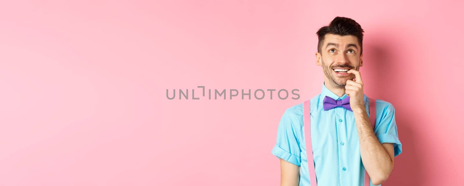 Image of smiling man making choice, looking dreamy and happy up, biting fingernail with tempted expression, wanting something, standing over pink background.