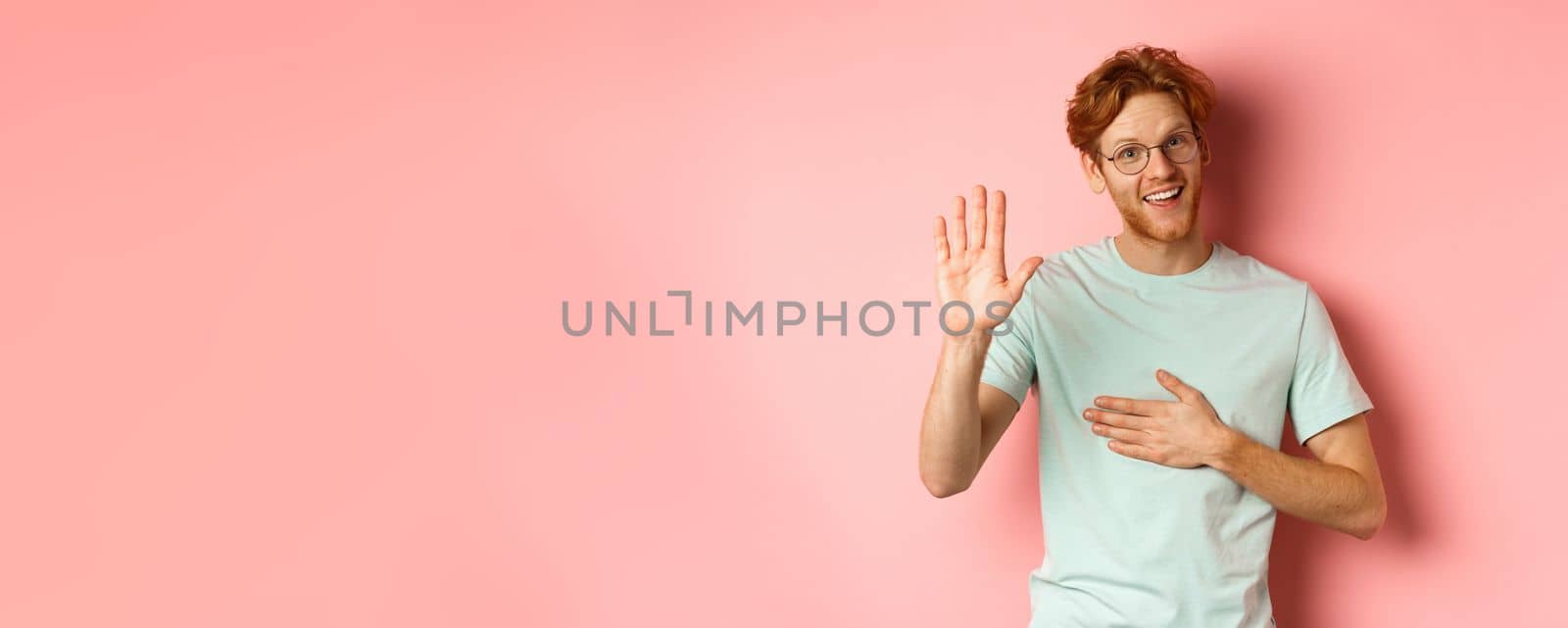 Friendly redhead man being honest, holding hand on heart and arm raised high to swear or make promise, smiling at camera, telling truth over pink background.