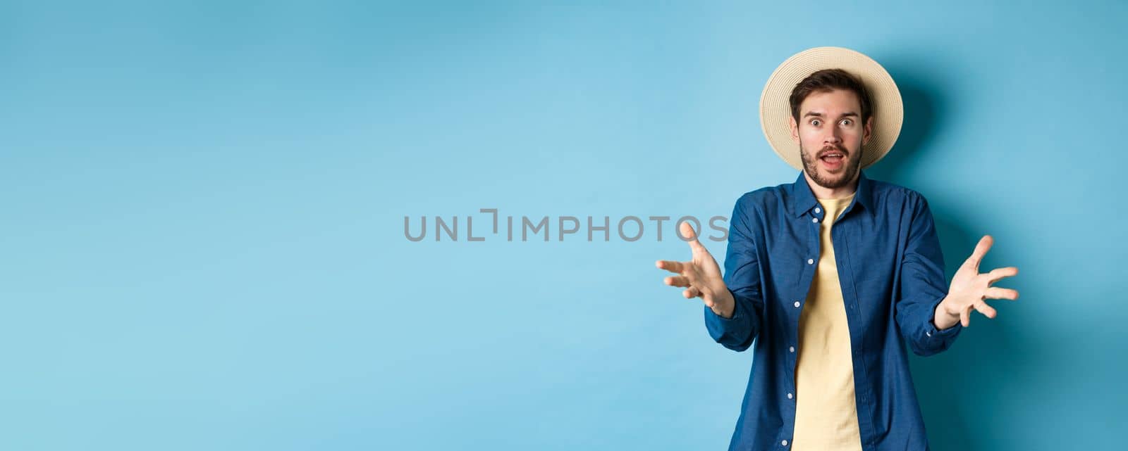 Surprised young man in summer hat, raising hands up staring with disbelief and amazement at camera, impressed with big event, standing on blue background by Benzoix